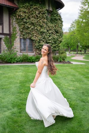 Smiling bride in strapless white gown twirling on green lawn outside ivy-covered building.