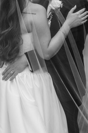 Black and white photo of bride and groom embracing, showing wedding dress, veil, ring, and boutonniere.