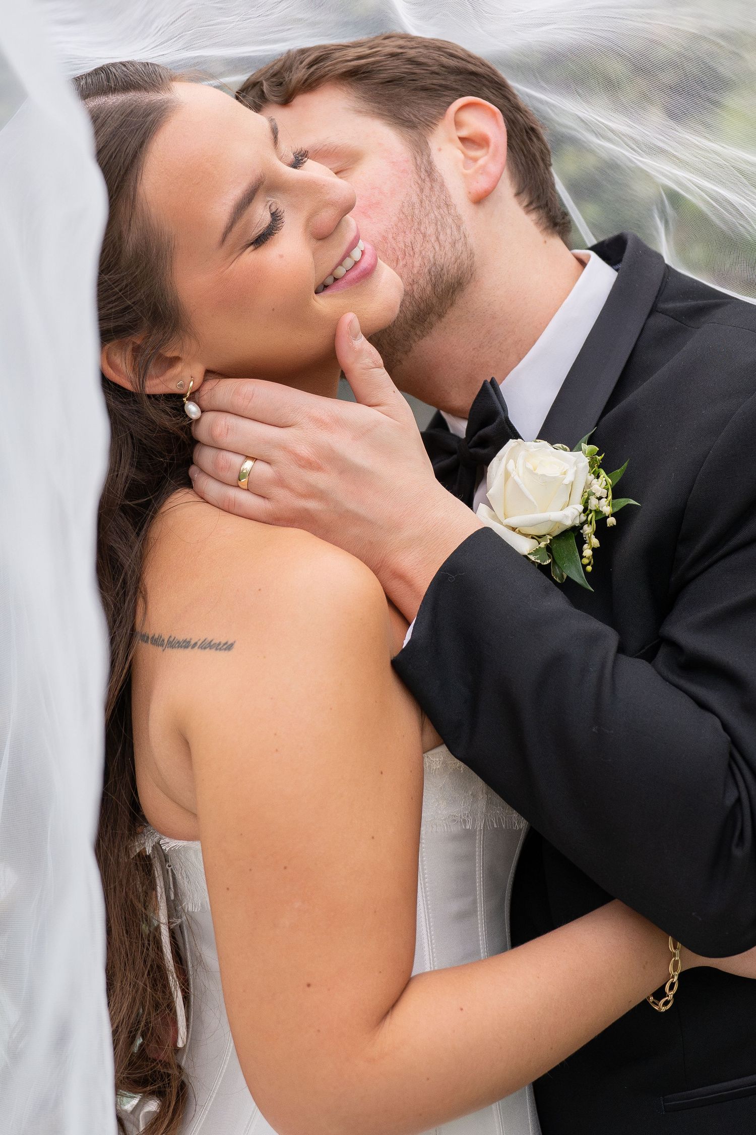 Groom in black tuxedo kissing bride on cheek under veil, bride smiling with eyes closed on wedding day.