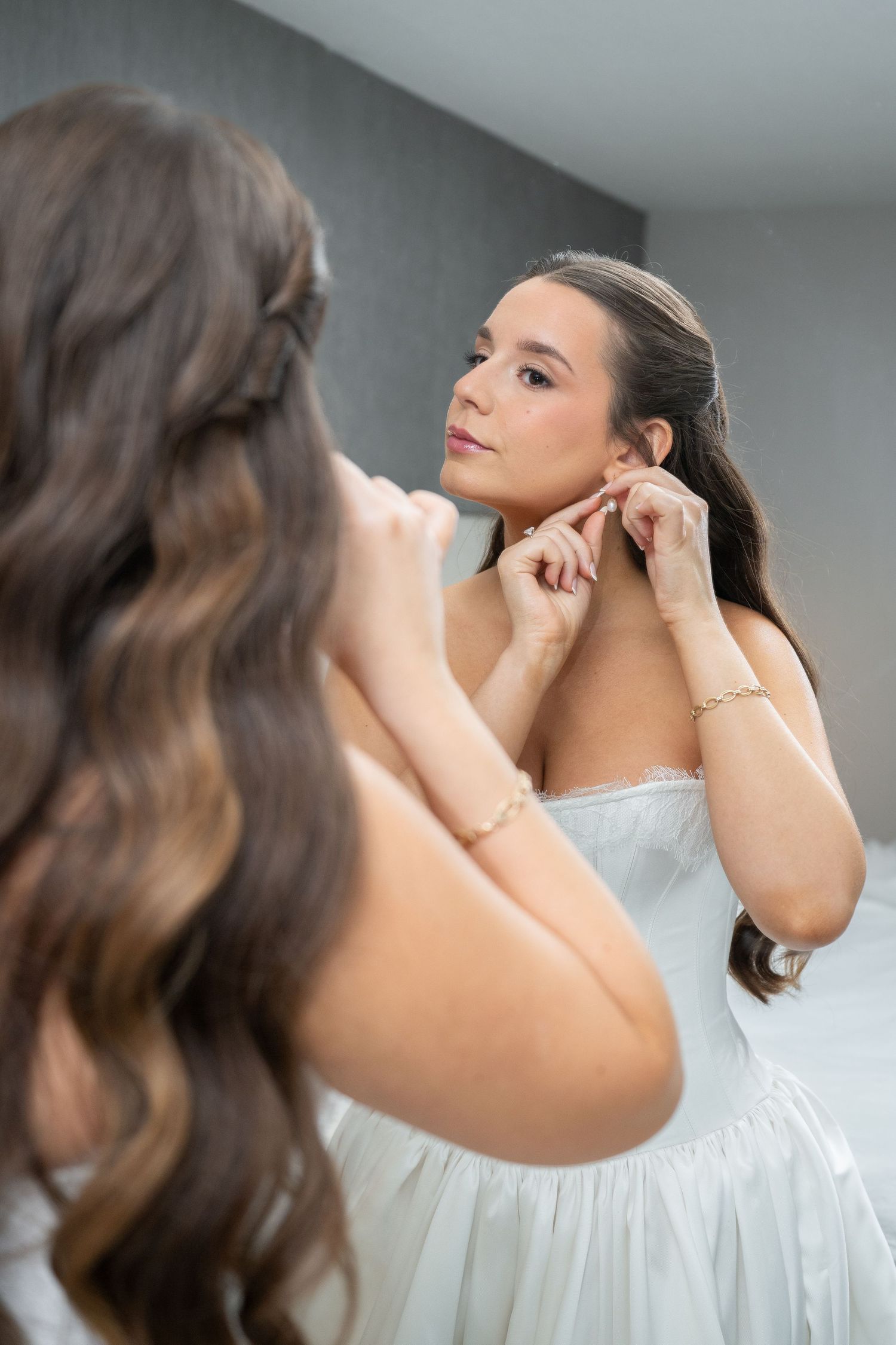 Bride in white strapless dress putting on earrings while looking in mirror during wedding preparation.