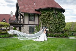 Bride and groom kissing on green lawn with flowing cathedral veil, Tudor-style mansion backdrop.