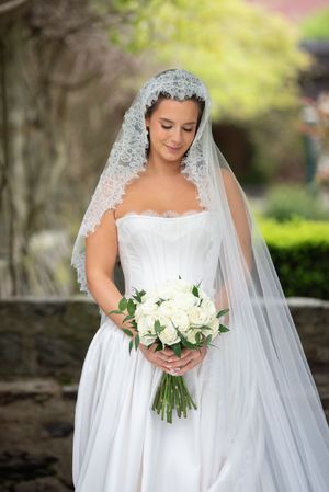 Bride in strapless white gown and lace veil holding white rose bouquet, standing outdoors in garden setting.