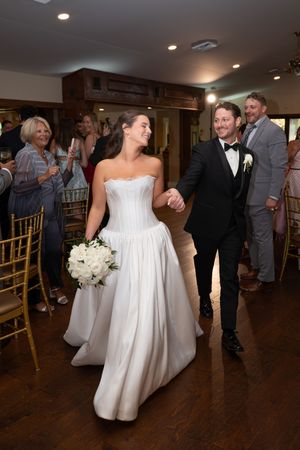 Bride in white strapless gown holds bouquet while walking with groom in black tuxedo at indoor wedding reception.