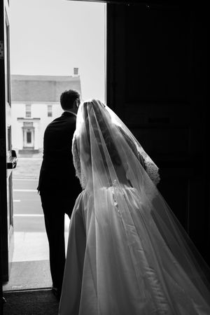 Black and white photo of bride in veil and groom in tuxedo standing at church doorway on wedding day.