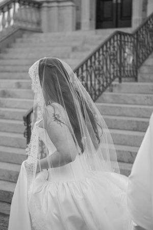 Black and white photo of a bride in a wedding gown and veil ascending stone stairs with ornate railings.