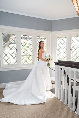 Bride in strapless white gown with long train holds bouquet by diamond-pane windows on a sunlit staircase landing.