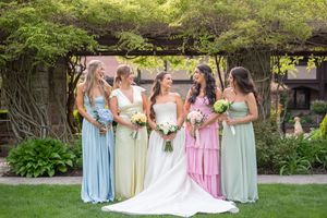 Bride in white gown with four bridesmaids in pastel dresses posing outdoors in a lush garden setting.