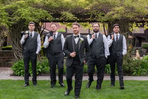 Groom and groomsmen in dark suits and vests pose confidently outdoors at a garden wedding venue.