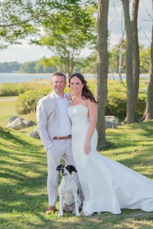 Newlyweds in white attire pose with their dogs under trees near waterfront.