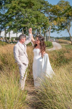 Couple in white attire dancing playfully among tall grass on sunny day.