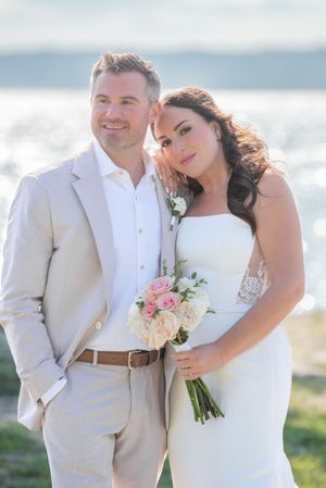 Wedding couple in light colored attire pose together on beach with floral bouquet.