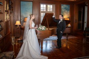 Elegant wedding couple stands in a grand wood-paneled room with natural light streaming through tall windows.