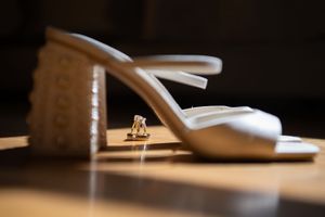 White wedding shoes and perfume bottles cast dramatic shadows on a wooden surface in natural light.