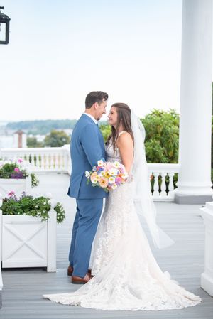 A wedding couple shares a romantic moment on a white columned porch with colorful bouquet.