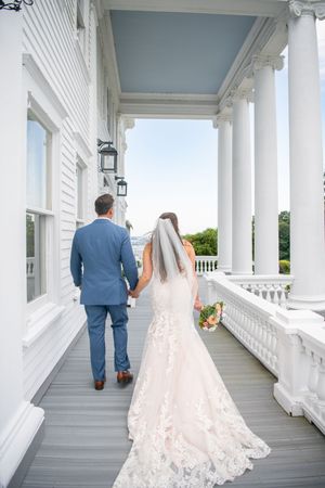 A couple walks along an elegant white columned porch with a trailing lace wedding dress.