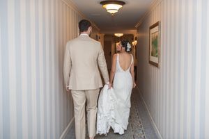 A couple walks down a hallway with striped wallpaper, showcasing the back of an elegant white wedding dress.