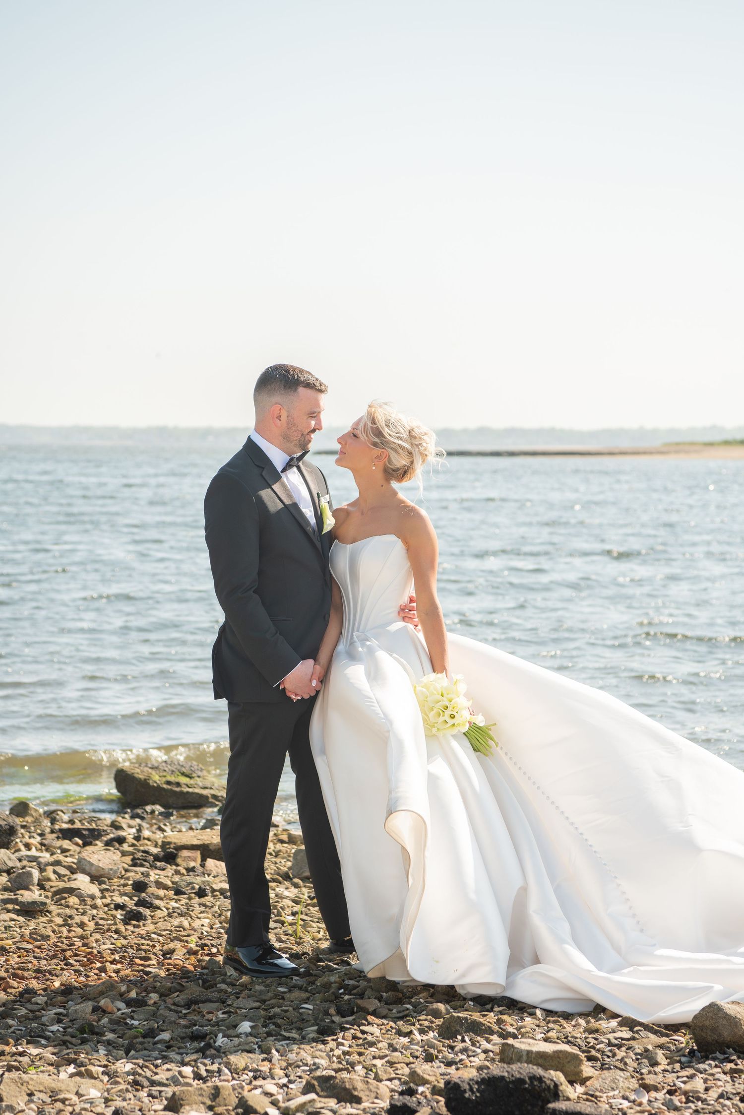 Elegant wedding portraits on a rocky beach shoreline with flowing white gown catching the breeze.