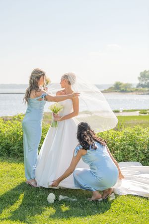 Bridesmaids in light blue dresses help arrange the bride's wedding gown on a sunny lawn.