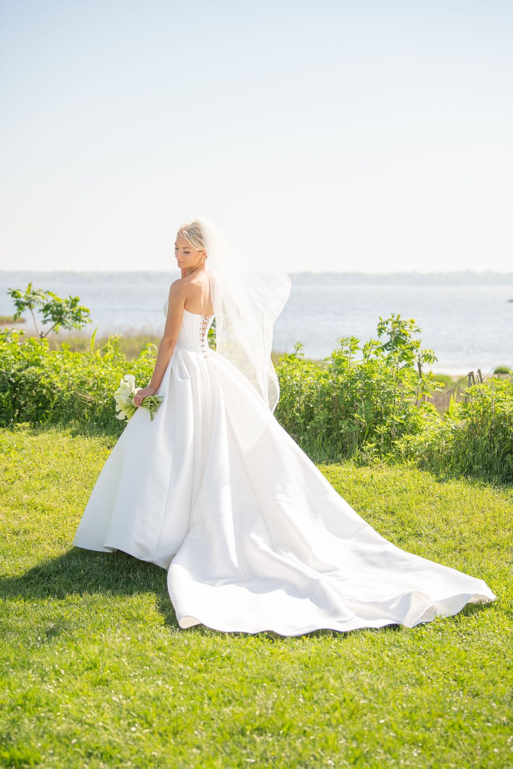 Bride in flowing white gown poses gracefully on green oceanfront lawn during golden hour.