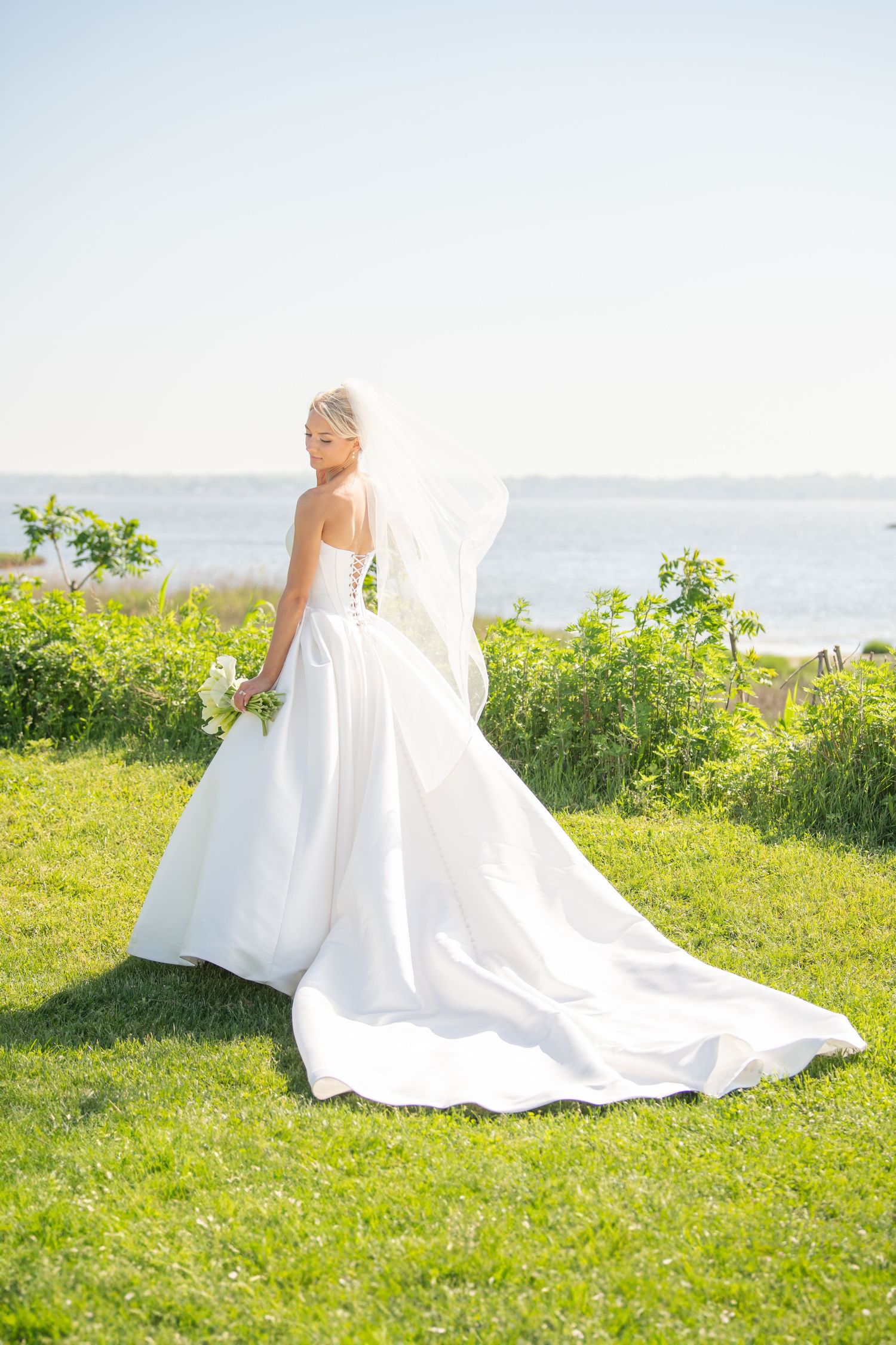 Bride in flowing white gown poses gracefully on green oceanfront lawn during golden hour.