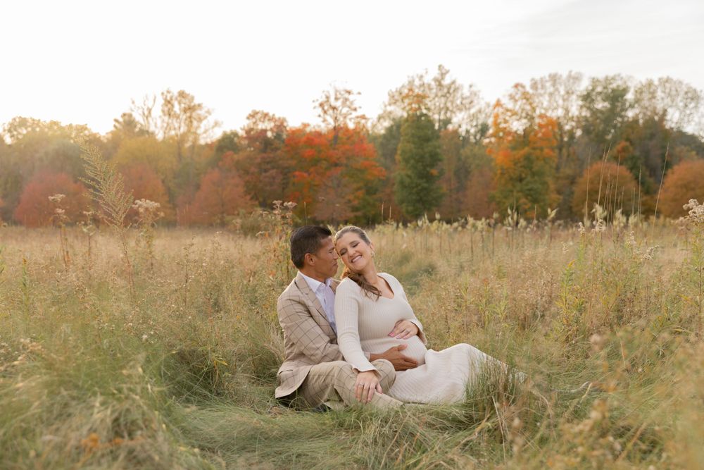 man in a tan suit sitting in a field with his pregnant wife in the fall