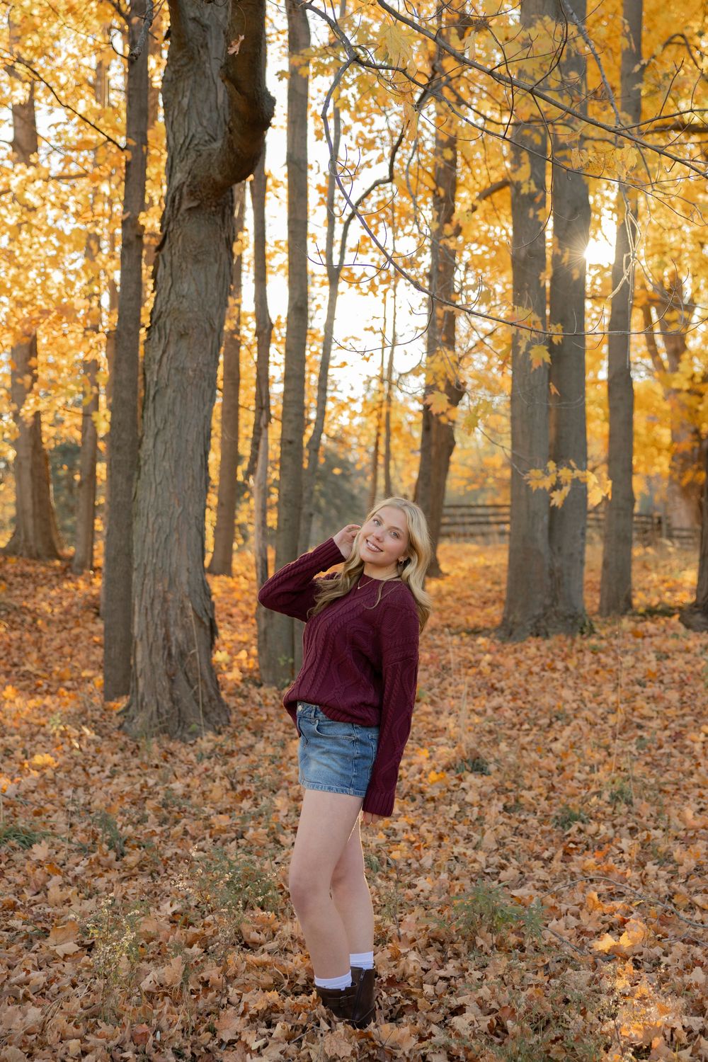 high school senior in a sweater poses among golden autumn trees with fallen leaves scattered in the forest in Rochester Hills michigan