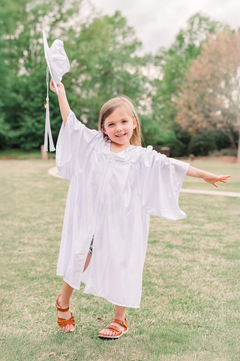 Kindergarten girl graduate in white cap and gown posing with a greenery background, Newnan, GA