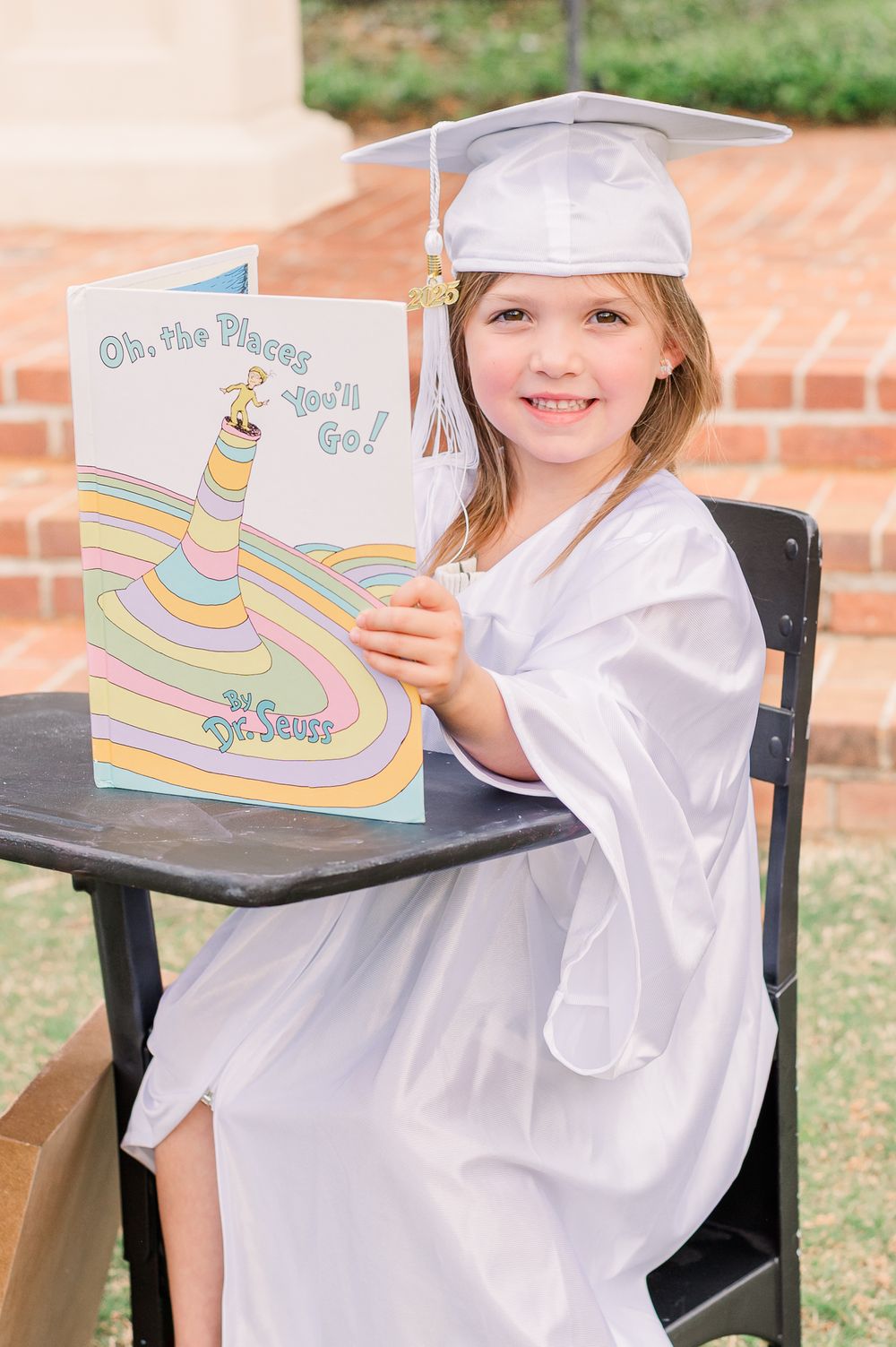 Kindergarten graduate in white cap and gown reading at a desk, Newnan, GA