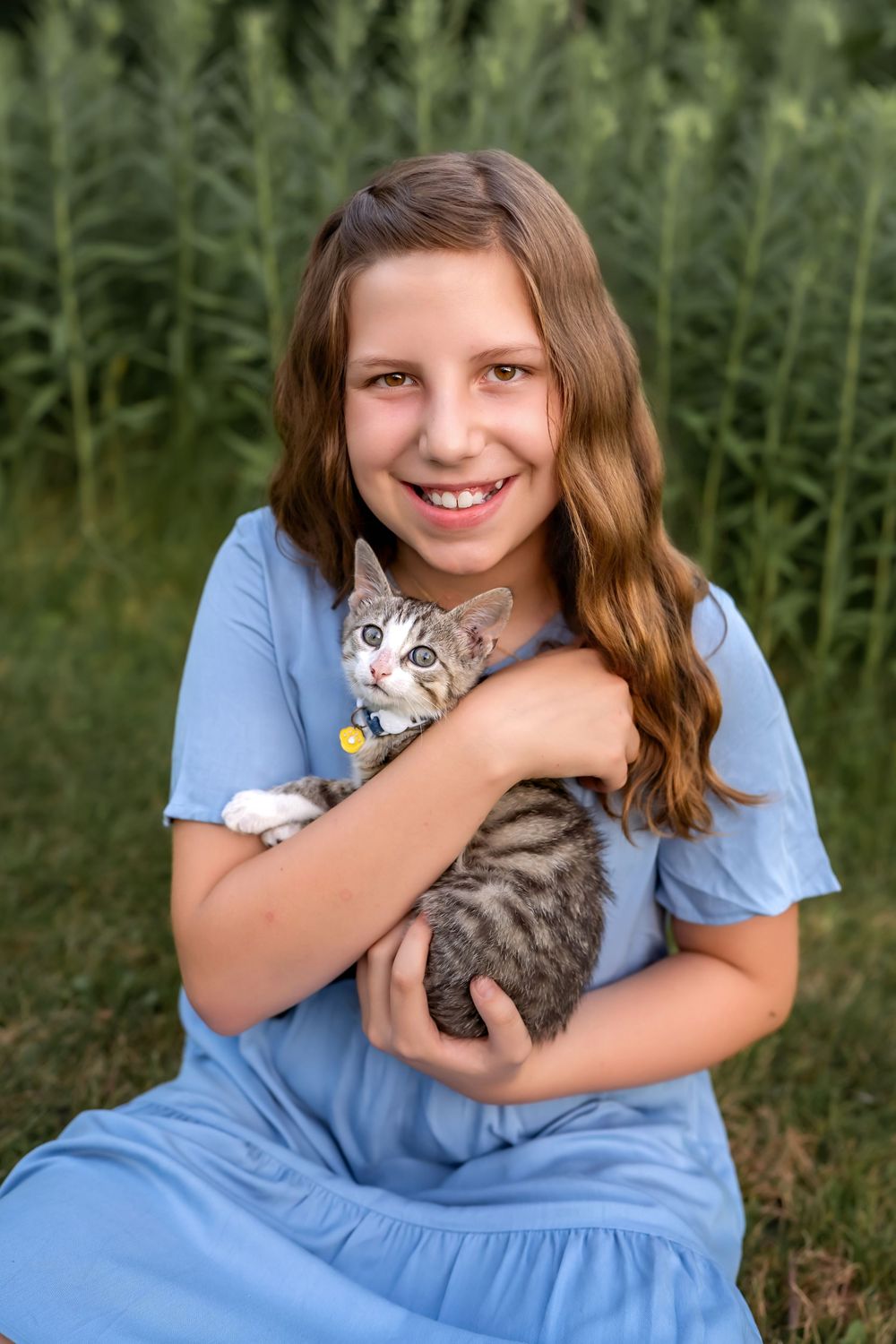 Young person in blue dress holds and cuddles adorable gray tabby kitten outdoors among green plants.