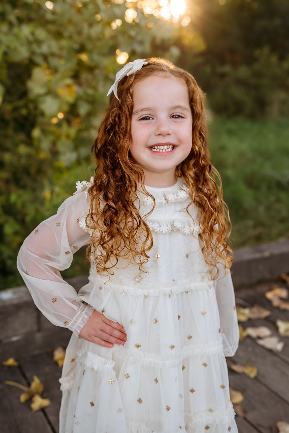 Child wearing a delicate white dress with gold accents poses outdoors during golden hour.