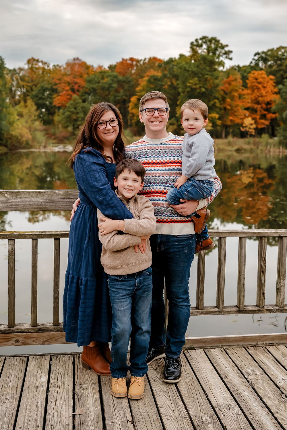 A family poses together on a wooden dock during autumn with colorful fall foliage and a lake in the background.