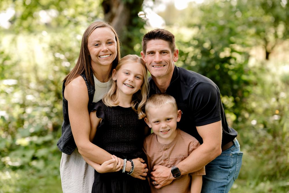 Happy family of four smiling outdoors in a lush green garden setting for a professional portrait.