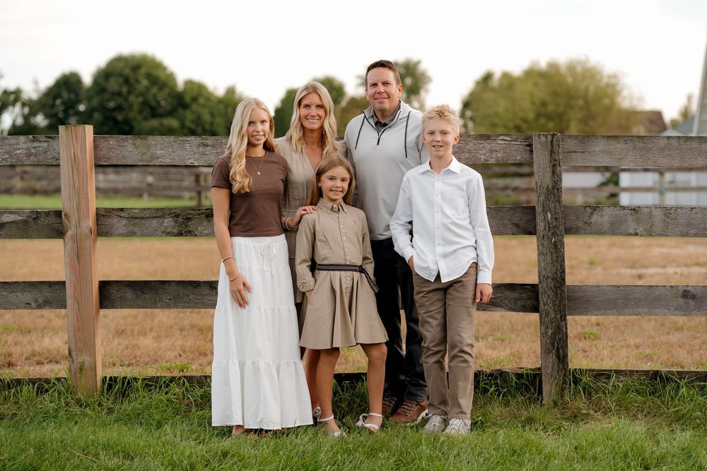 Family of five smiling together outdoors by a wooden fence on a green lawn during golden hour.