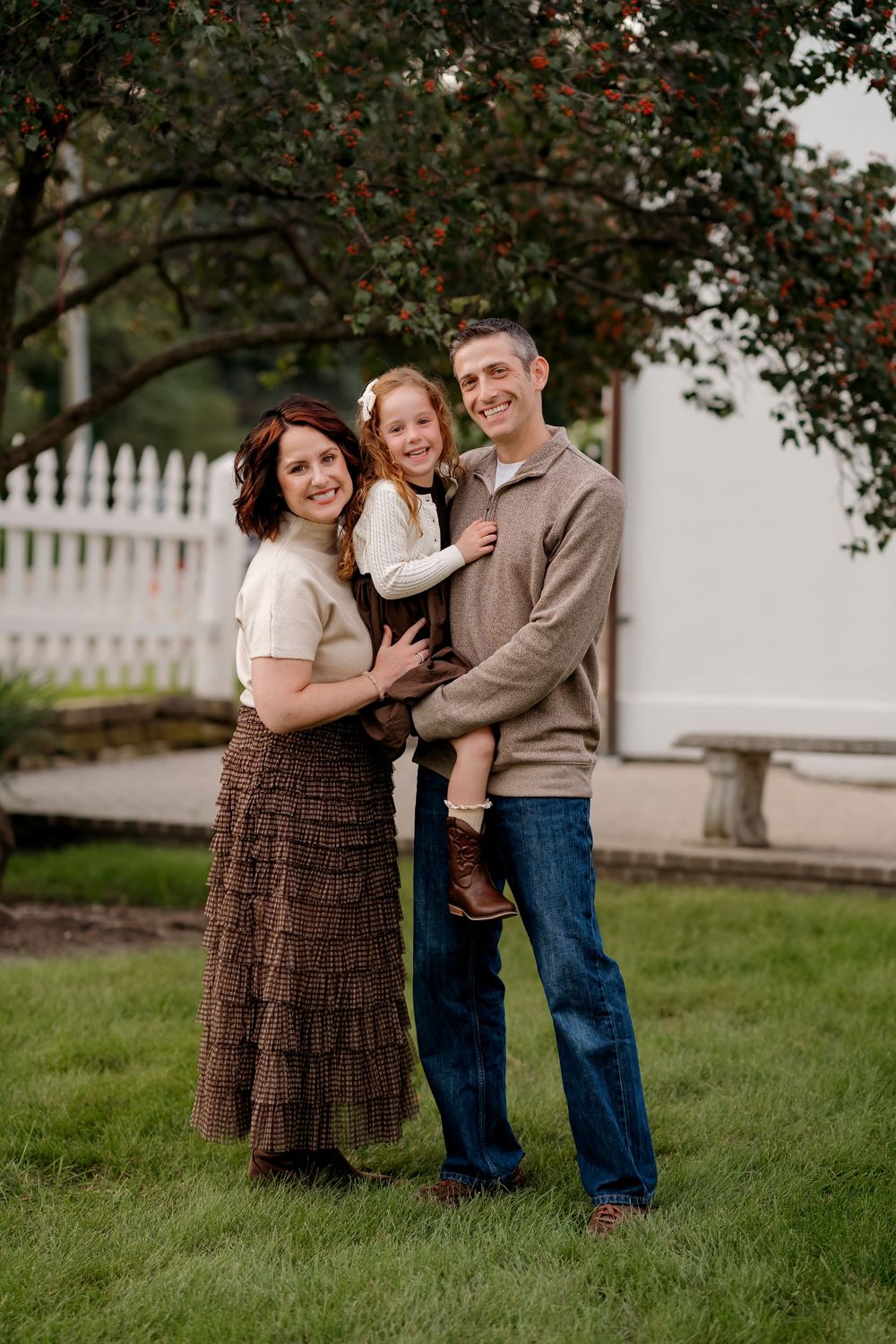 A smiling family of three poses outdoors on green grass, with a white picket fence and berry tree behind them.