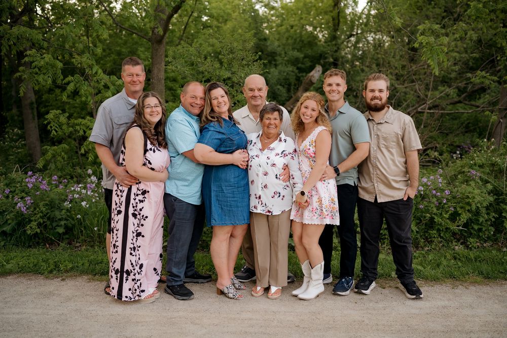 A joyful family group of nine poses together outdoors on a path surrounded by lush green trees and purple wildflowers.
