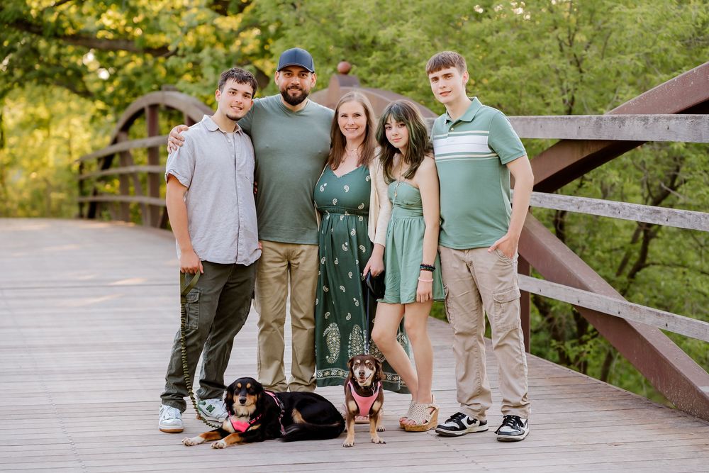 Family of five poses on a wooden bridge with two dachshunds, surrounded by lush green trees.