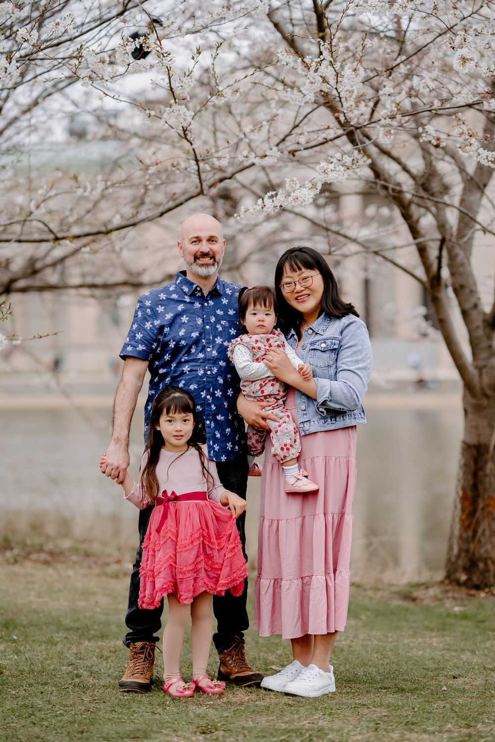 Family of four posing under blooming cherry blossom trees near a waterway, wearing coordinating pink and blue outfits.
