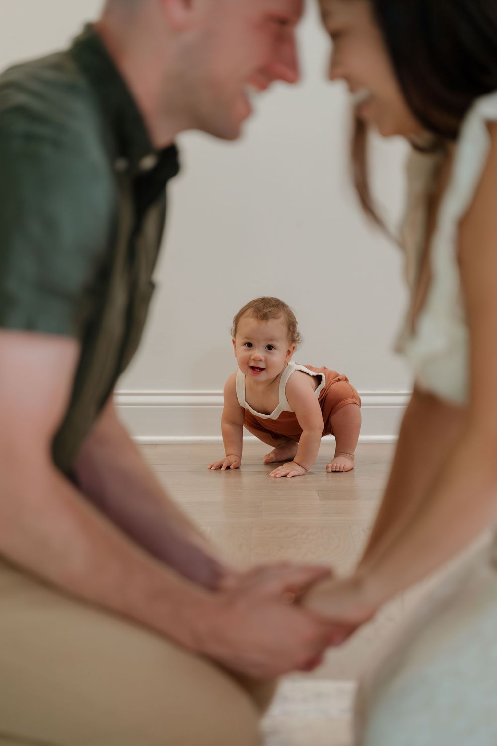 A happy baby crawls on hardwood floor while two parents lean in smiling during a warm family portrait session.