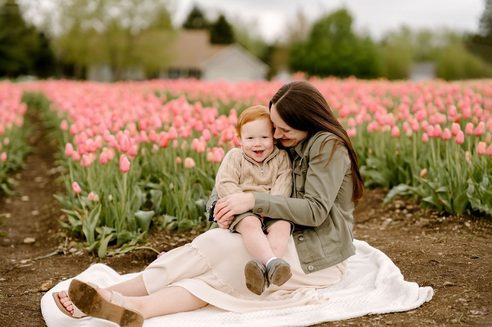 Mother and toddler sitting on blanket in pink tulip field, sharing a tender moment outdoors.