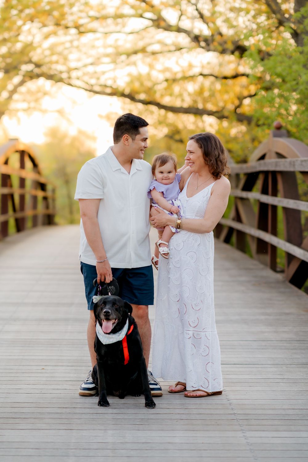 Family portrait on a wooden bridge at sunset with a man, woman holding baby, and a black Labrador dog.