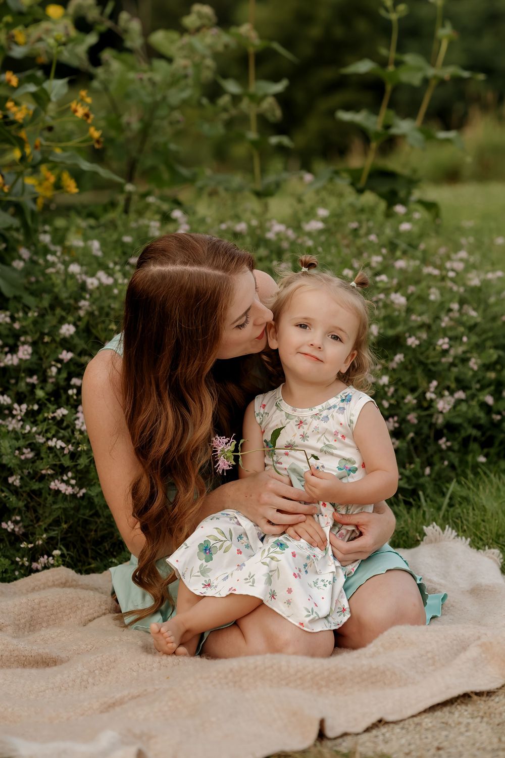 A mother kisses her toddler daughter on the cheek while sitting on a blanket in a lush garden.