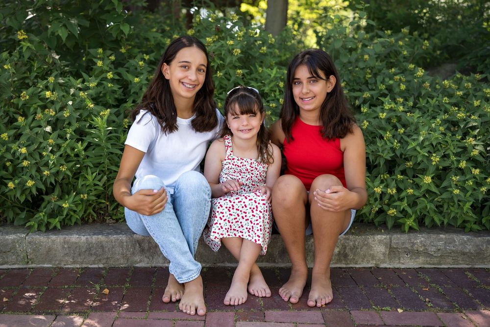 Three people sit together on outdoor concrete steps surrounded by green foliage in summer.