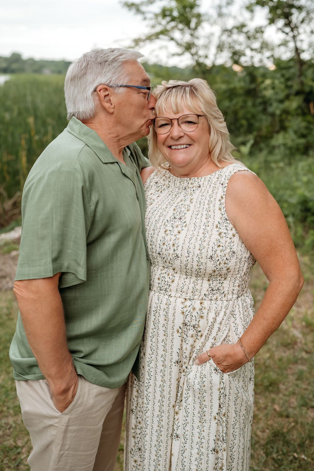 An affectionate mature couple poses outdoors, with the woman in a white dress and man in green polo shirt.