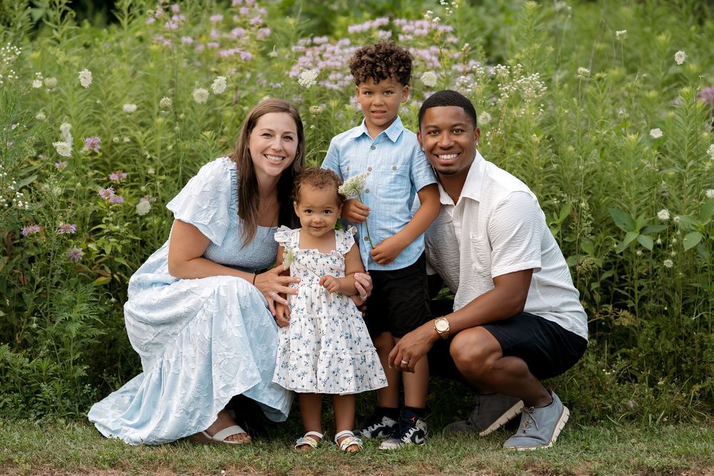 Family of four smiling outdoors in a wildflower garden, parents with two young children posing for a portrait.