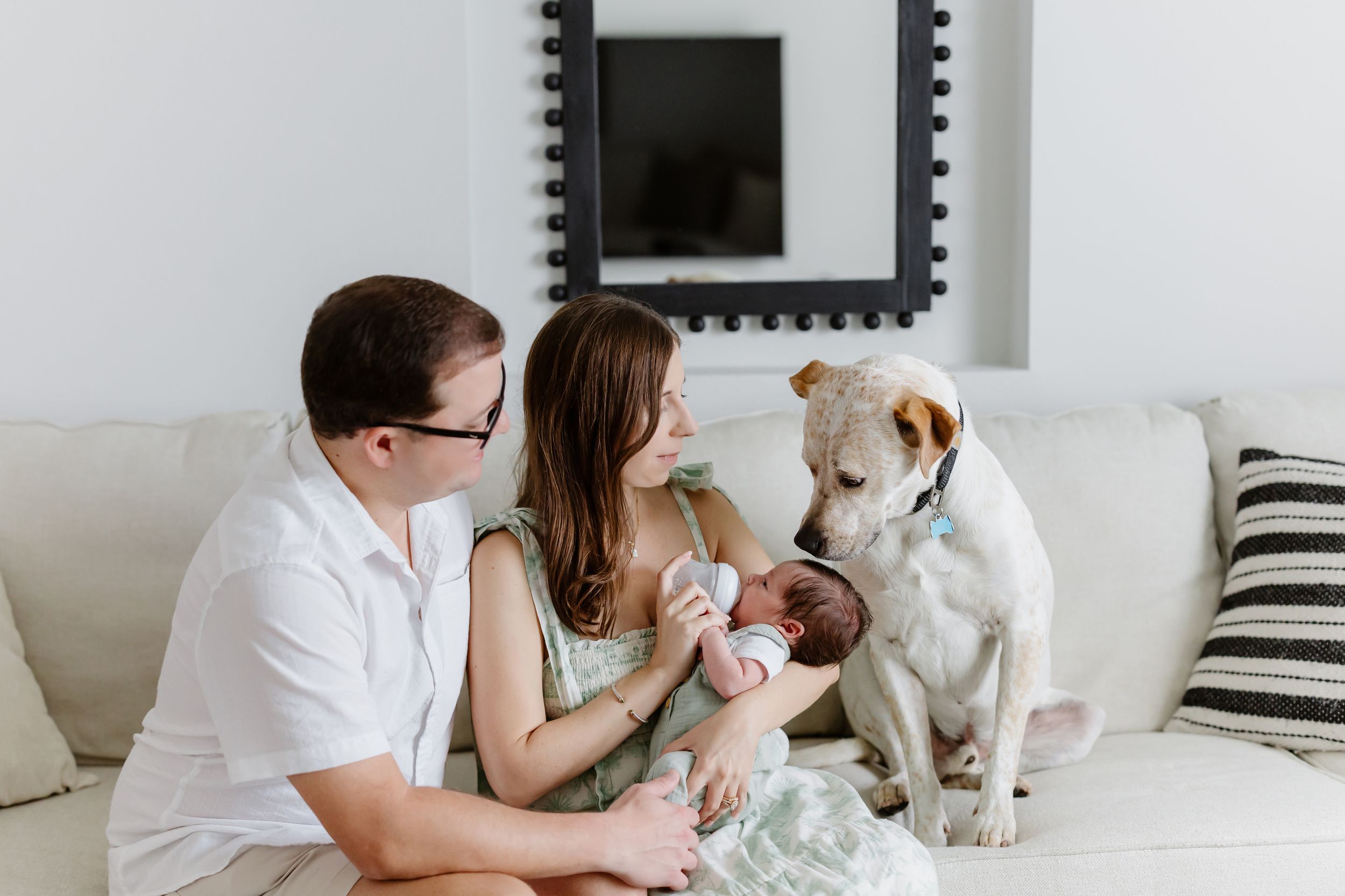 Family relaxes on white couch with their dog and newborn baby in bright modern living room.