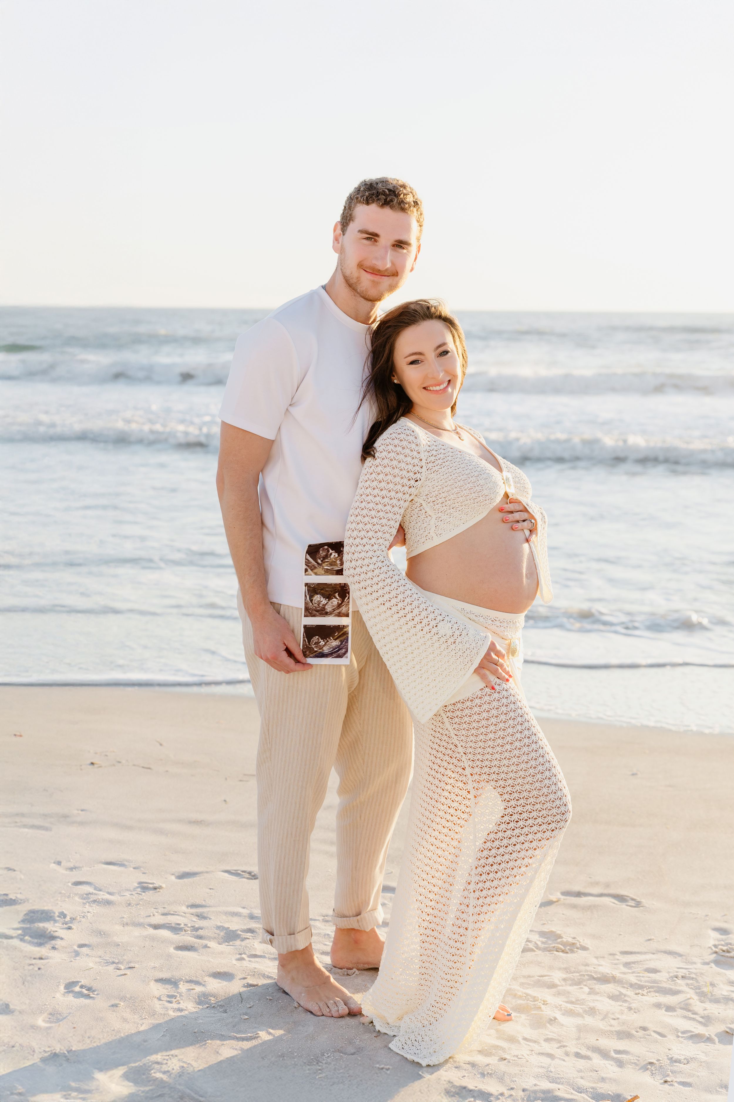 A couple embraces on a sunny beach while holding an ultrasound photo during a pregnancy announcement photoshoot.