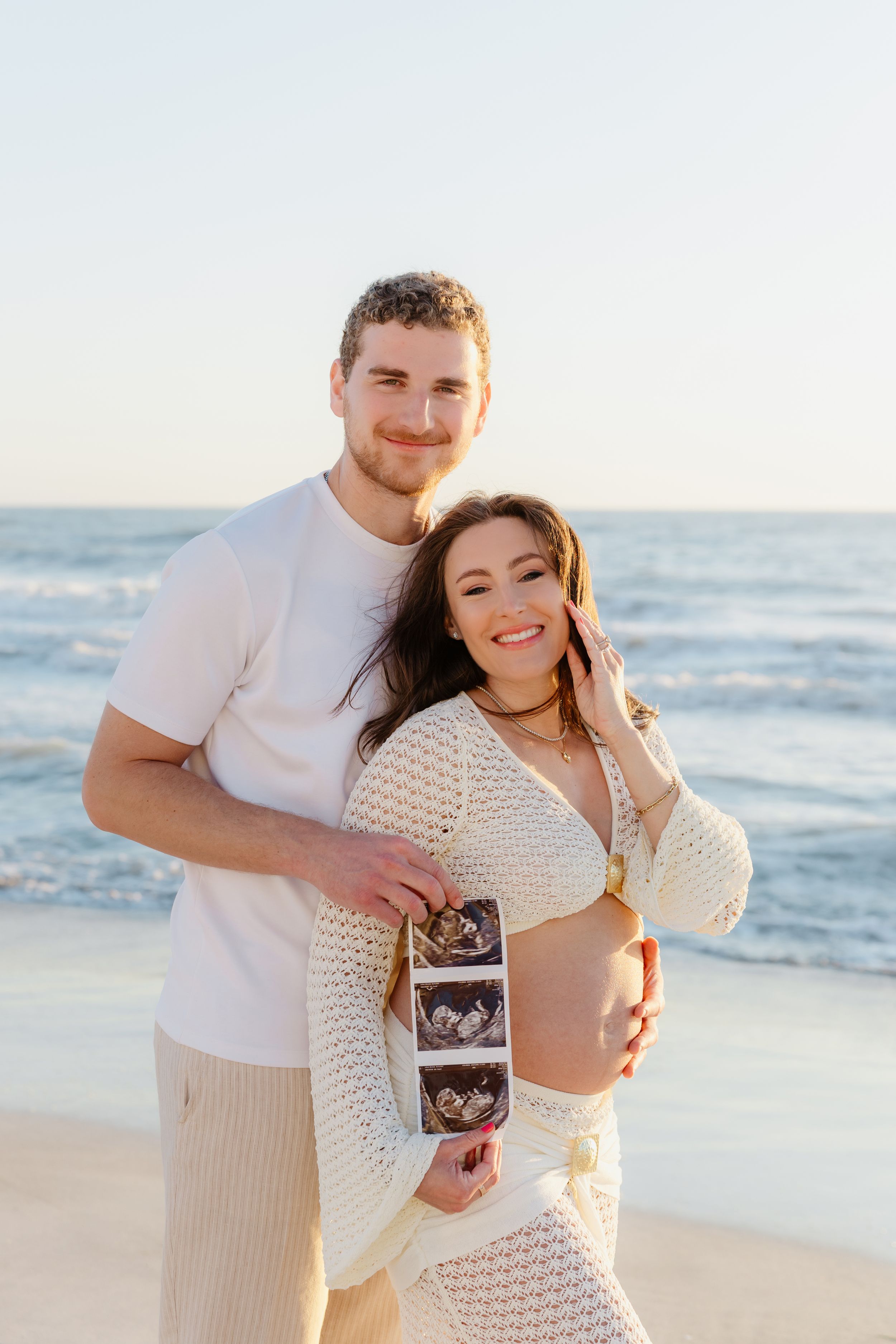 A couple shares an intimate moment on the beach while holding a sonogram photo during sunset.