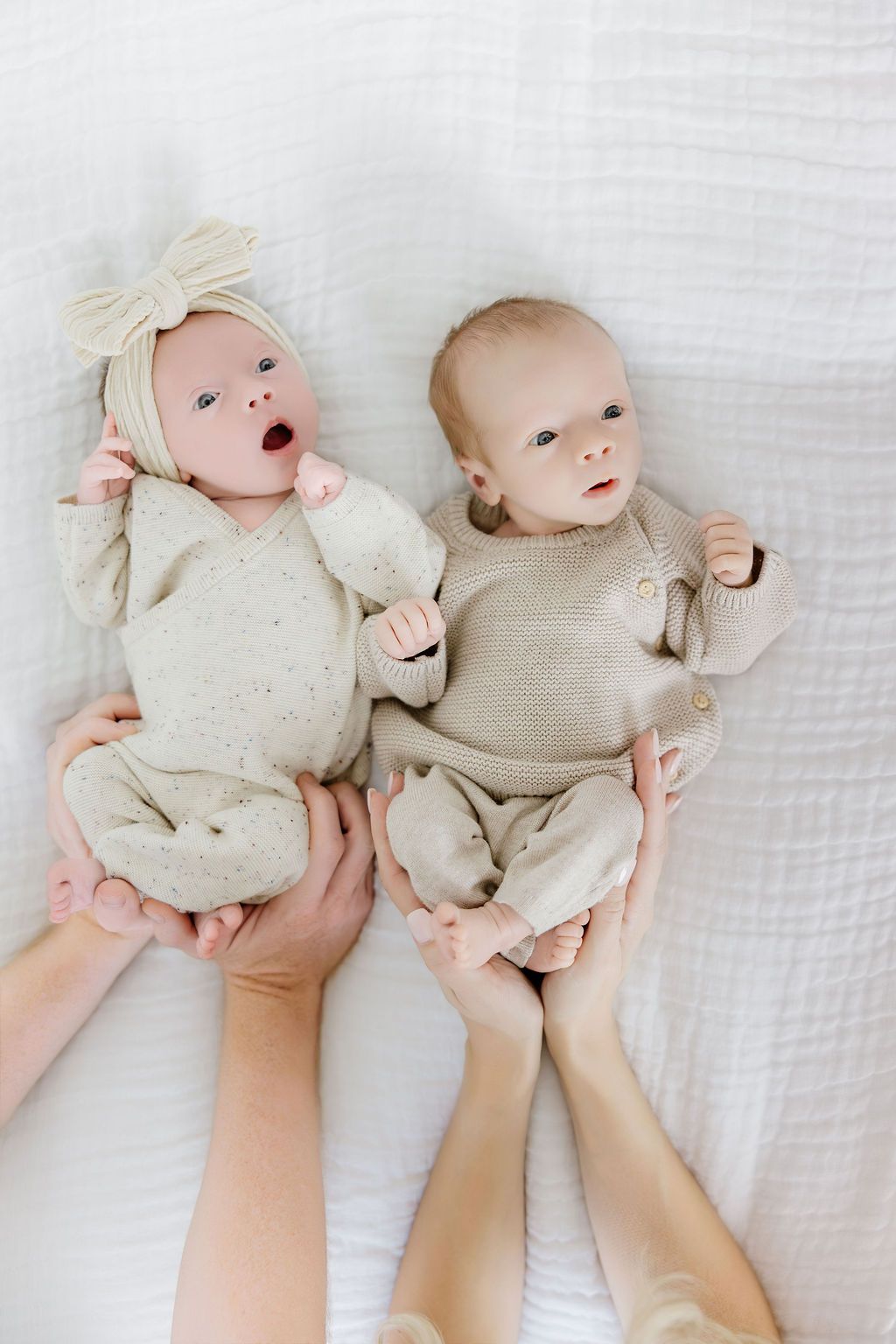 Two newborn twins in beige knit outfits lying on white fabric, gently held by adult hands.