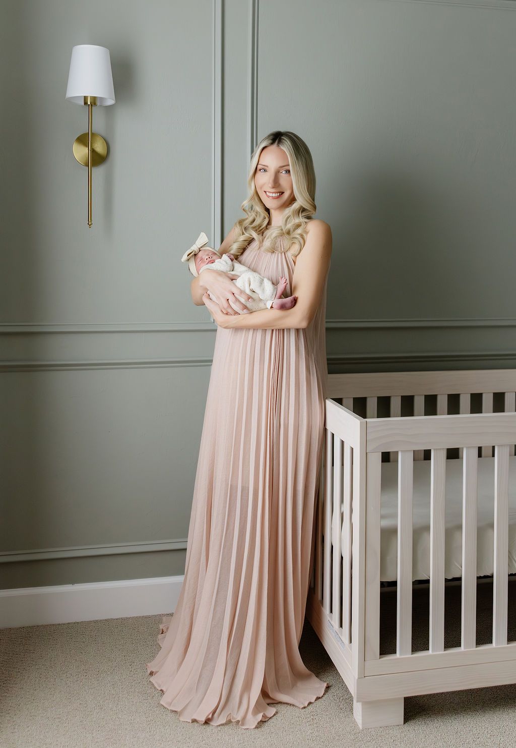 Mother in blush pleated gown holds newborn baby girl in elegant sage green nursery with white crib.