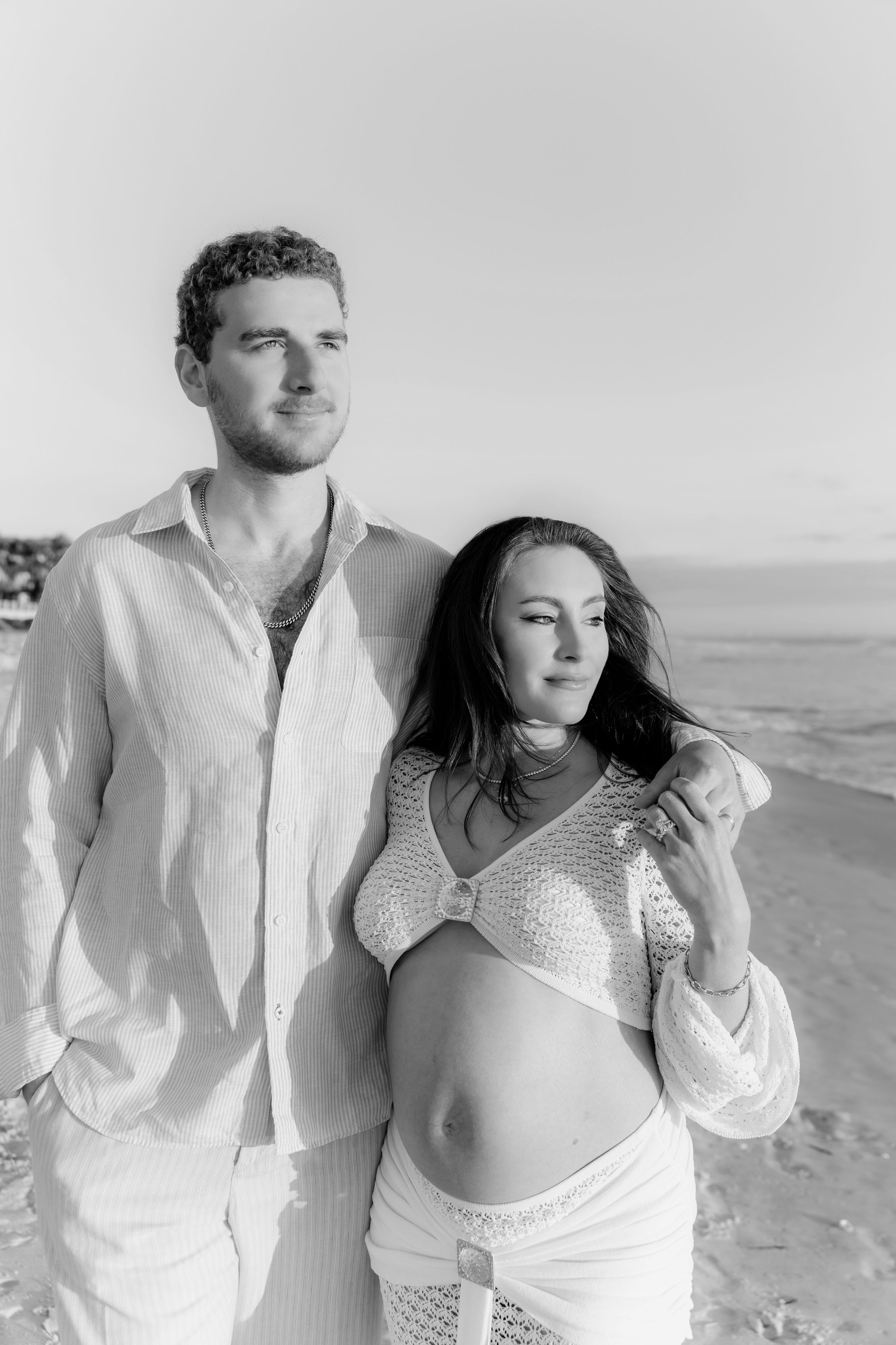 A couple embraces on a sandy beach in white outfits while displaying a sonogram photo.